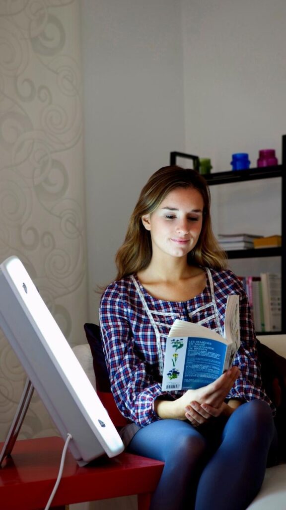 young woman reading book by light therapy lamp