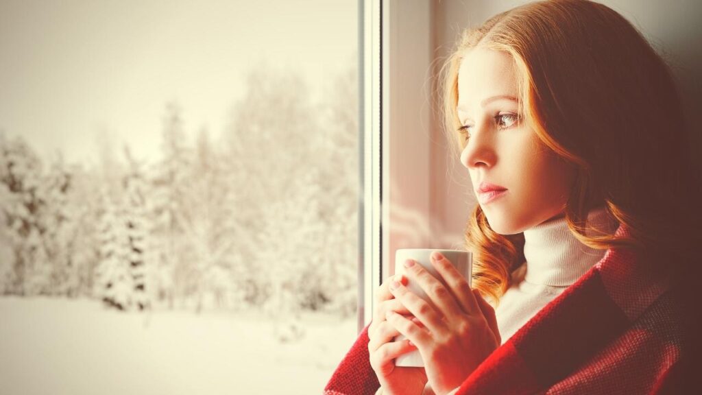 sad girl holding hot drink and looking out at snow